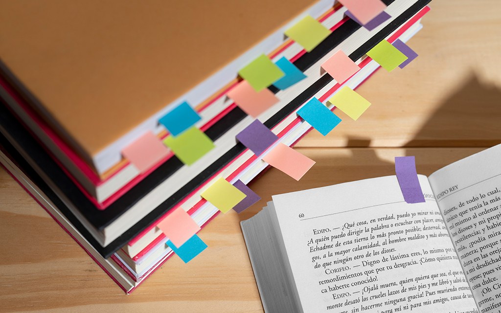 Stack of books with colorful sticky tabs next to an open book on a wooden table.